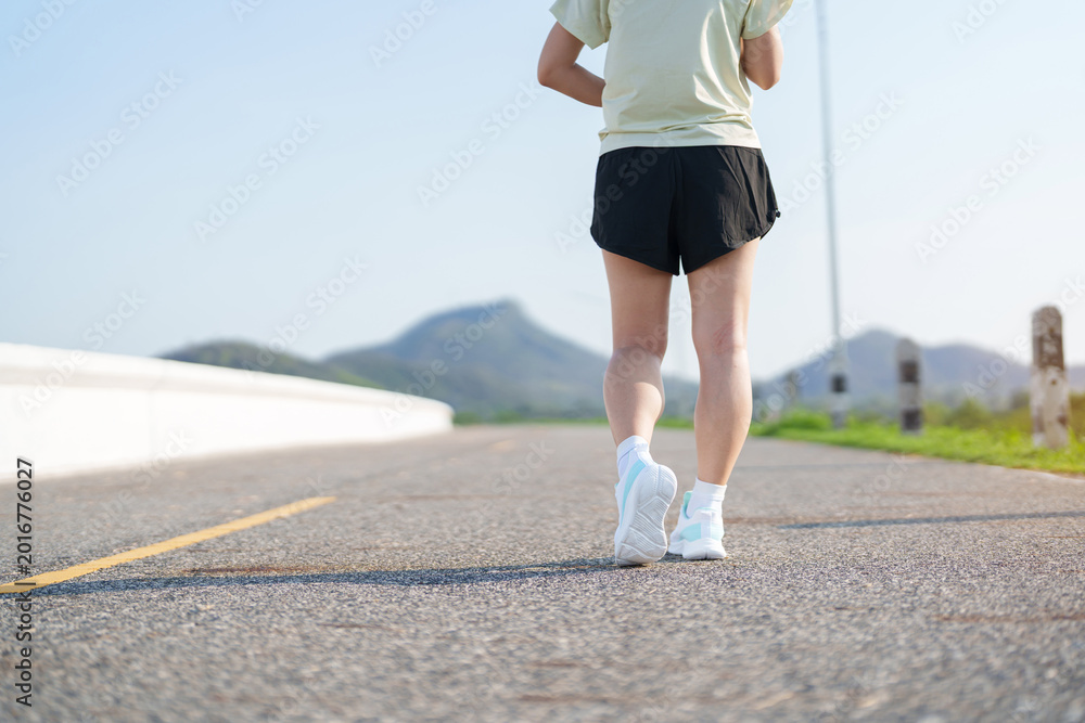 Fototapeta premium Young adult Asian woman jogging and walking in the park at morning. Female athlete in sport shoes running on the road with visible leg muscles. Healthy lifestyle, wellness and workout concept