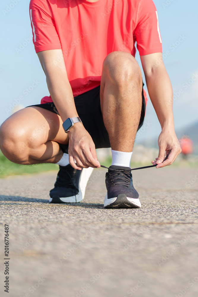 Fototapeta premium Young Asian man tying running shoes in the park. Female runner getting ready for jogging and exercise on the road. Fitness, wellness and outdoor sport concept in morning