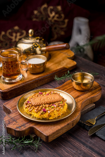 Photography kunafa topped with lotus jam, chopped pistachios, edible roses and lotus biscuits with sugar water served on a gold plate on a wooden coaster