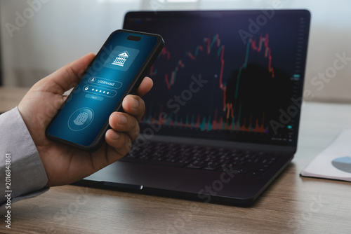 Hand holding smartphone with mobile online banking login screen in front of laptop displaying financial trading chart, suitable for fintech security, digital payment, and online transaction concepts.