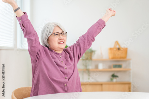 Фотография A smiling senior woman raises her arms in a victory pose in her living room (feeling refreshed, invigorated, happy, accomplished, successful)