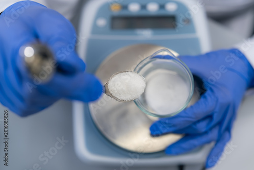 Weighing a precise amount of white crystalline substance using a stainless steel spoon over a glass beaker at an analytical balance station.