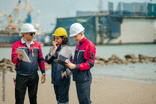 Logistics engineer writing operational report while maritime team communicates through radio system during industrial cargo port inspection and shipping management process.