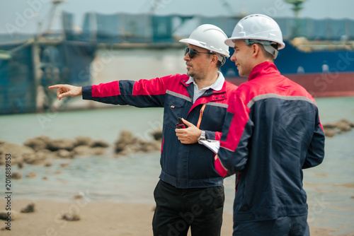 Maritime engineering manager pointing toward industrial shipping zone while discussing cargo port inspection and logistics planning with harbor operations team member.
