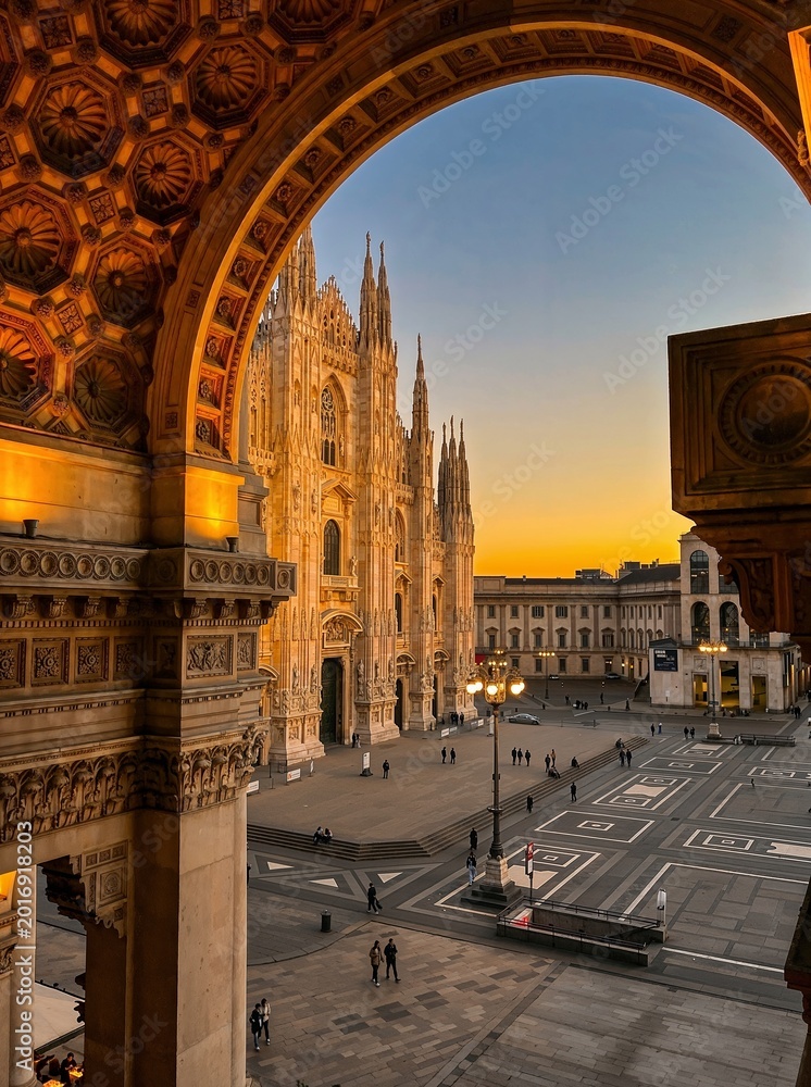 Fototapeta premium Golden sunset light frames the majestic Gothic cathedral through ornate arches in Milan, creating a timeless architectural scene filled with elegance, symmetry, and historic European charm.