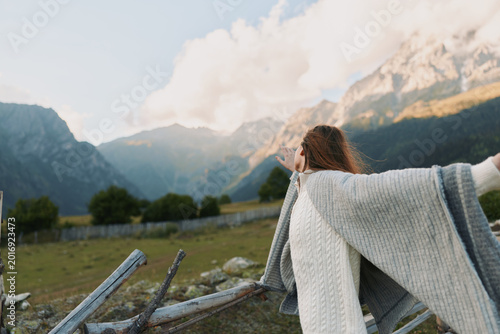 Fotografi Young woman with curly hair wrapped in a cozy knitted shawl enjoys fresh mountain air while standing near a wooden fence in a serene rural landscape