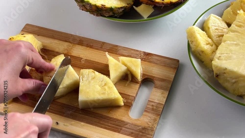 A woman's hands slice a fresh pineapple into pieces and trim away the tough, inedible part of the fruit on a wooden cutting board.