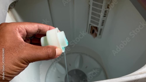 Close-up of a person pouring blue liquid detergent into a top-loading washing machine.