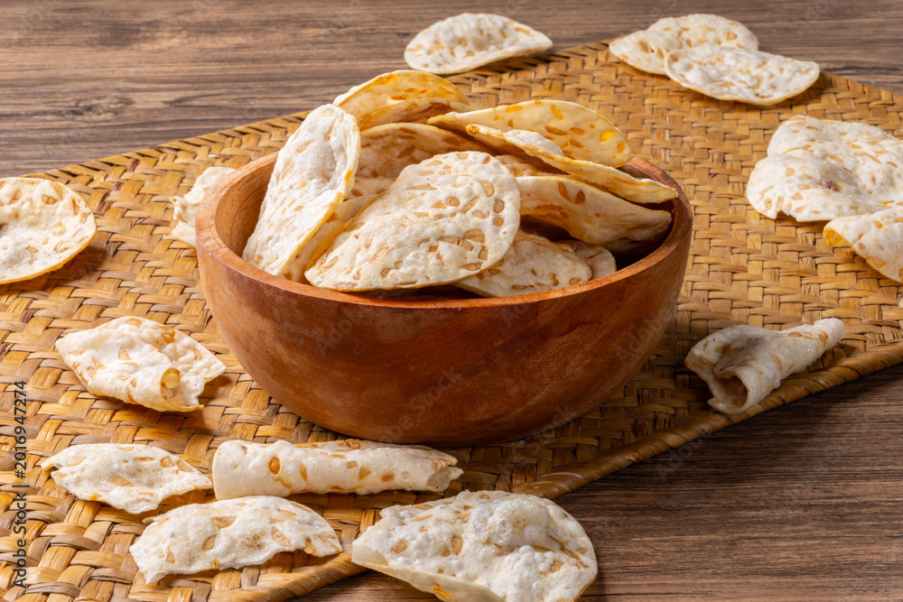 Fototapeta premium Crispy tempeh chips arranged in a wooden bowl on a traditional woven mat and rustic table