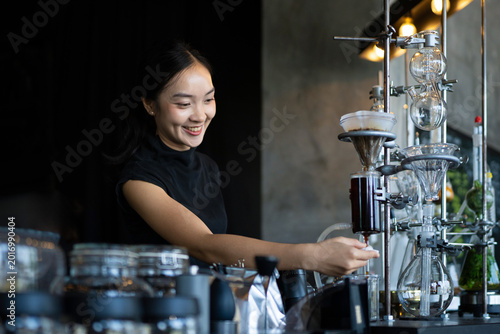 Schilderij op canvas Young Asian woman barista smiling while operating an elegant siphon coffee brewi