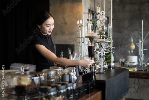 Tableau sur toile Young Asian woman barista smiling while operating an elegant siphon coffee brewi