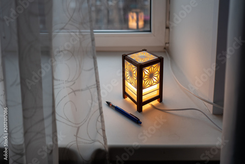Bild auf Leinwand A warm wooden Kumiko lantern glows on a window sill beside a pen, casting soft ambient light and creating a cozy, calm evening atmosphere ideal for reading, writing or quiet reflection
