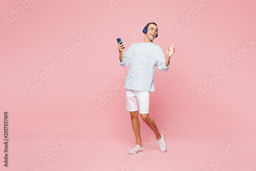 Full body young middle eastern man he wear casual blue cotton shirt listen to music in headphones use mobile cell phone isolated on plain pastel light pink wall background studio. Lifestyle concept