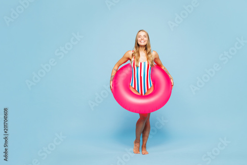 Full body young smiling satisfied woman she wears bikini swimsuit rest near hotel pool hold in hand pink rubber ring look camera isolated on plain blue background. Summer vacation sea sun tan concept.