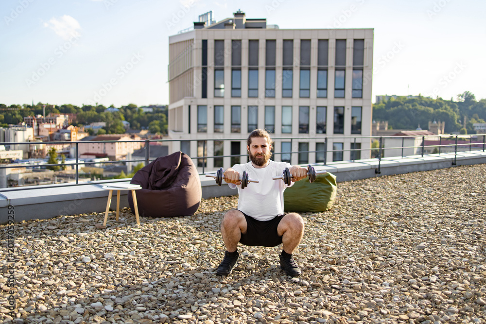 Fototapeta premium A bearded man with long hair performs dumbbell squats on a rooftop terrace with a city view and beanbag chairs