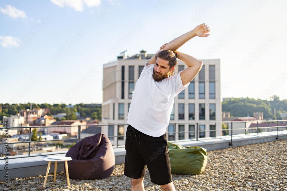 Fototapeta premium A man with a beard and bun hairstyle performs a shoulder stretch on a rooftop terrace with a city view and modern building in the background