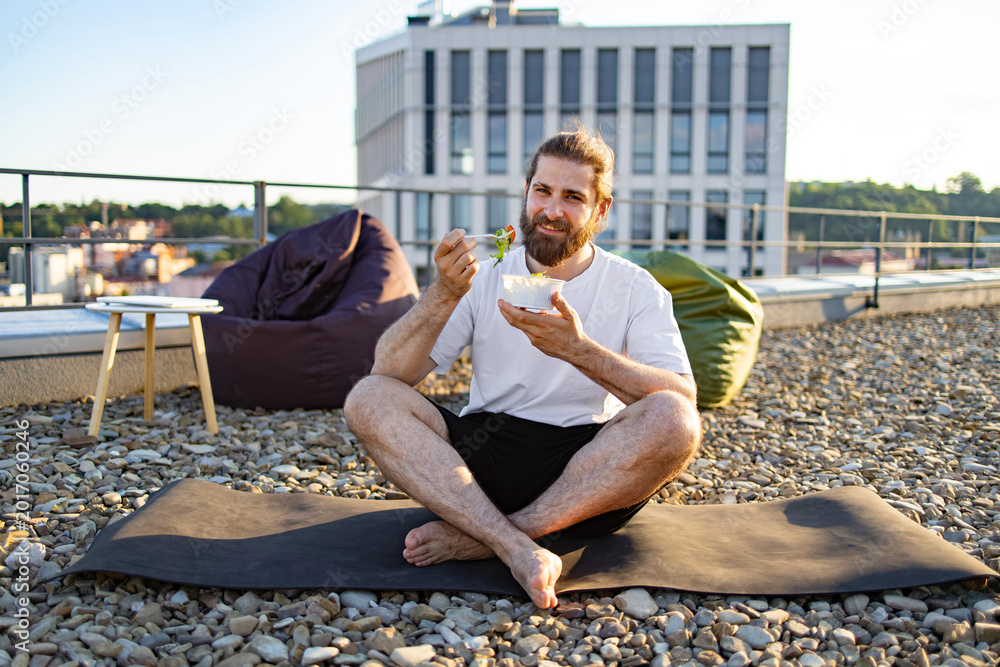 Naklejka premium A bearded man in casual attire enjoys a healthy salad while sitting cross-legged on a yoga mat on a gravel rooftop with a city view