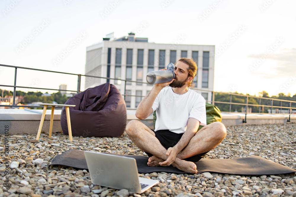 Naklejka premium A man with a beard and long hair sits cross-legged on a yoga mat on a gravel rooftop, drinking from a water bottle while a laptop is open in front of him