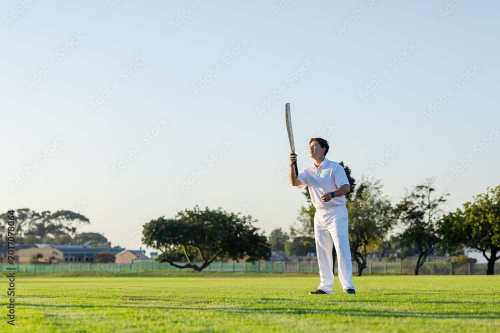 Obraz premium Male cricketer in whites standing on pitch holding bat, sporting wristband, looking up, copy space