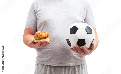 A fat male soccer fan in gray shorts and a t-shirt holds a soccer ball and a hamburger in his hand.