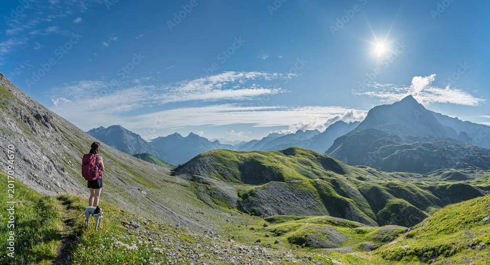 Fototapeta premium Epic mountain panorama with female hiker and dog looking at sun over alpine peaks, wide valley vista in Lechquellengebirge, Vorarlberg, Austria