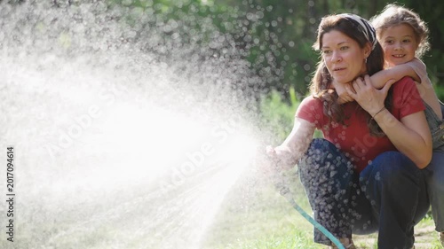 Young mother and her little daughter joyfully playing with a garden hose while watering the garden on a sunny summer day. Happy family bonding and carefree outdoor moments.