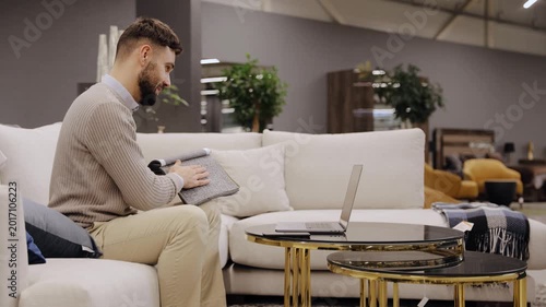 Man selecting sofa fabric samples, browsing options on laptop in modern furniture showroom