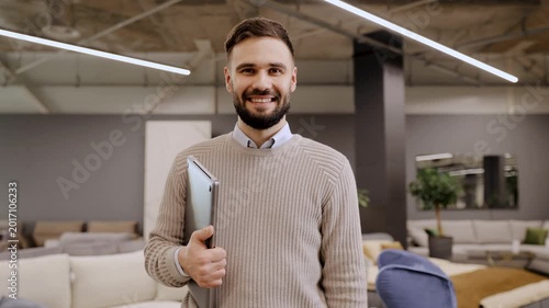 Smiling bearded man holding laptop, professional male in sweater looking at camera in modern office