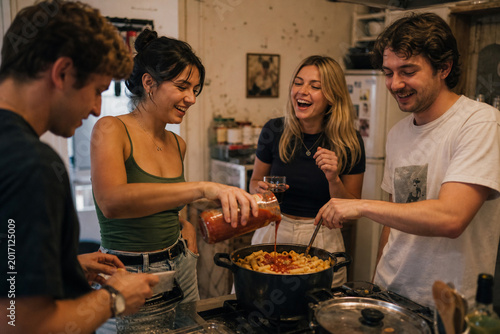 Four young adults cooking pasta and laughing together in a home kitchen, suitable for content about friendship, home cooking, and happy social gatherings.