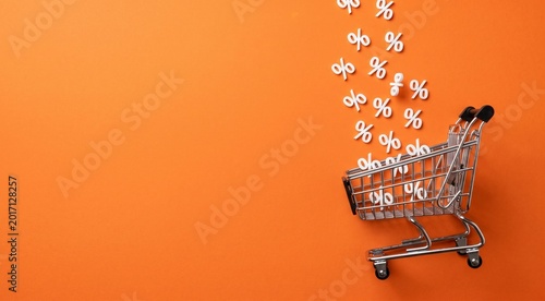 Minimalist shopping discount concept with white percentage symbols falling into a miniature metal shopping cart on a vibrant orange background, modern e-commerce sale theme