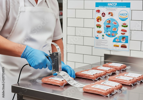 A worker in blue gloves applies labels to vacuum-packed sausages, ensuring proper food safety and quality control. A HACCP poster hangs nearby as a reminder of hazard analysis