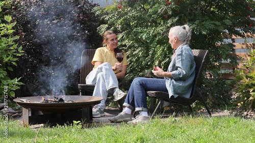 An elderly woman and her adult daughter drink wine and talk sitting by the fireplace in the garden.