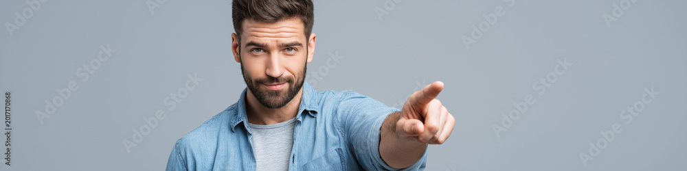 Obraz premium A man with a beard wearing a denim shirt points his finger forward with a serious expression against a neutral gray backdrop.