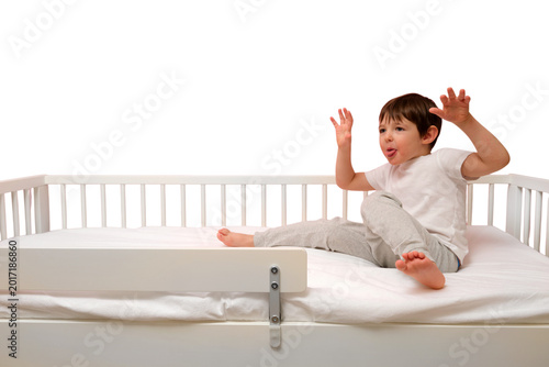 Child playing on bed with safety rail. Little boy sitting on mattress and making funny face with tongue out isolated on transparent background, interior furniture for bedroom for kid.