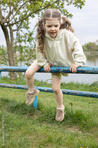 Happy Little Girl Climbing Sports Pipe on Playground Near Lake – Active Child Enjoying Outdoor Fitness and Nature
