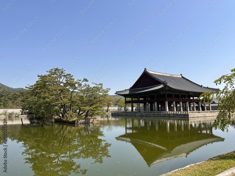 Naklejka premium Bright day by a calm lake with a traditional building reflecting in the water and trees surrounding the area in a historical park