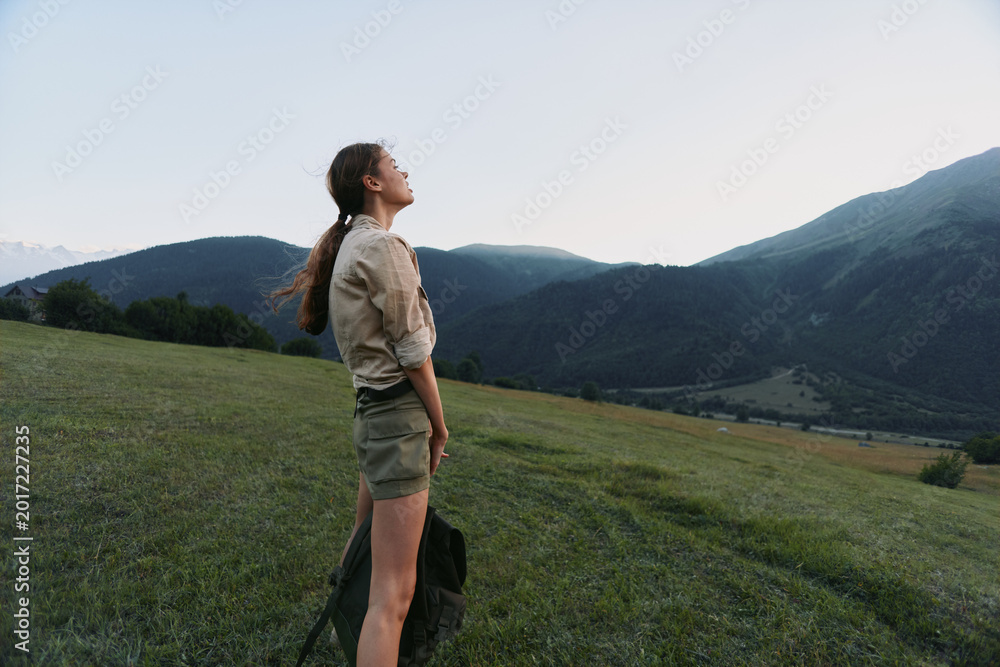 Naklejka premium Cheerful woman outdoors, standing in a sunlit meadow with distant mountains, wearing casual shorts and shirt, enjoying fresh air, a peaceful moment, and a relaxed lifestyle