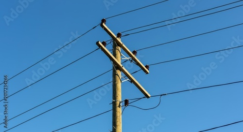 A massive wooden utility pole and electrical distribution network supporting vital wires, photographed from a low angle against a vast, bright, clear blue sky, transmission, network, electricity