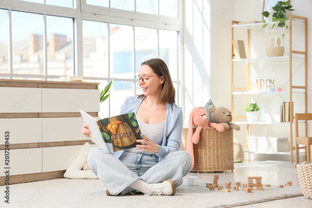 Fototapeta premium Female nursery teacher with book sitting on carpet in kindergarten