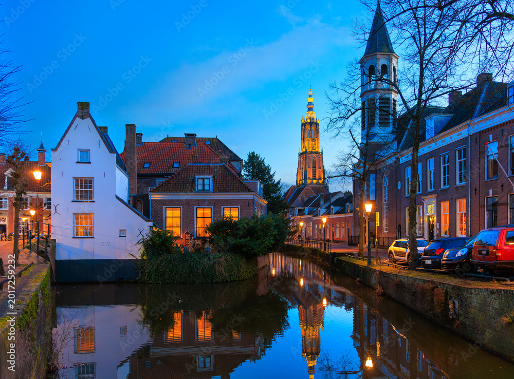 Fototapeta premium Dutch canal house with reflection in the canal, street lighting on, Onze Lieve Vrouwetoren clock tower in the background, Amersfoort, Netherlands, European lifestyle