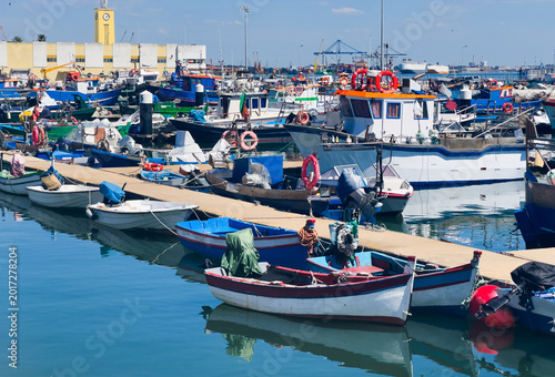 Photography Colorful traditional fishing boats moored in the calm, reflective water of Setubal harbor, Portugal