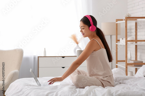 Beautiful young happy Asian woman in headphones with laptop and cup of tea li...