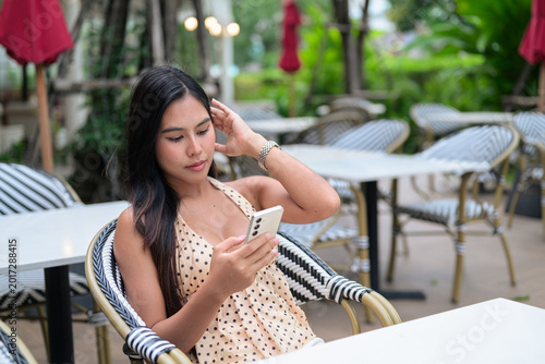 Beautiful Thai woman in a polka dot dress using a smartphone at an outdoor cafe during a leisure city travel break.