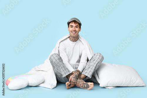 Tattooed young man in pajamas with pillow and blanket sitting on blue background