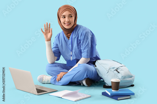 Female Muslim medical intern with laptop studying on blue background