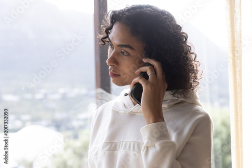 Non-binary person holding smartphone to ear, gazing out large window with sheer curtain, copy space