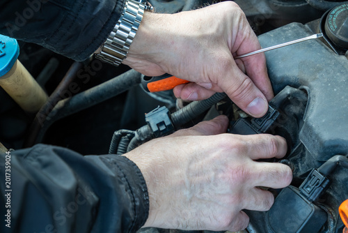 overhead view of male mechanic hands wearing metal watch using flat head screwdriver to pry plastic clip and disconnect electrical harness from black ignition coil pack