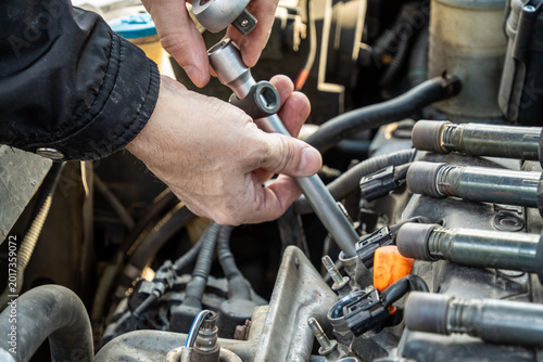 Foto male hands utilizing metal socket ratchet with extension to reach into deep engine cylinder wells for spark plug extraction while disconnected ignition coils lay nearby on top of car motor