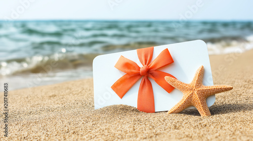 White gift card with ribbon and starfish on sandy beach with sea background. Concept of travel, vacation and holiday gift
