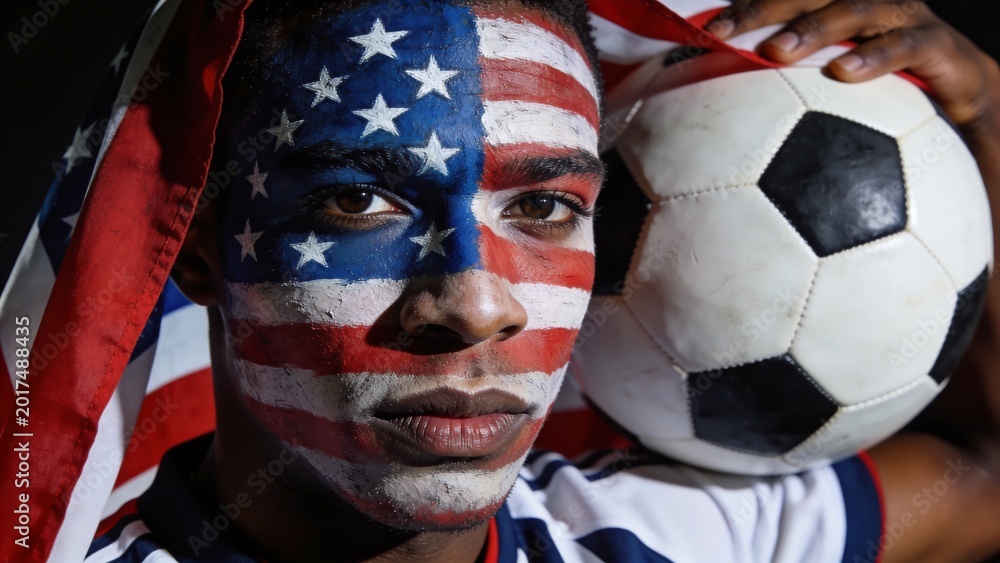 Fototapeta premium Face-painted man pressing black-white soccer ball to head in studio with US flag and striped jersey